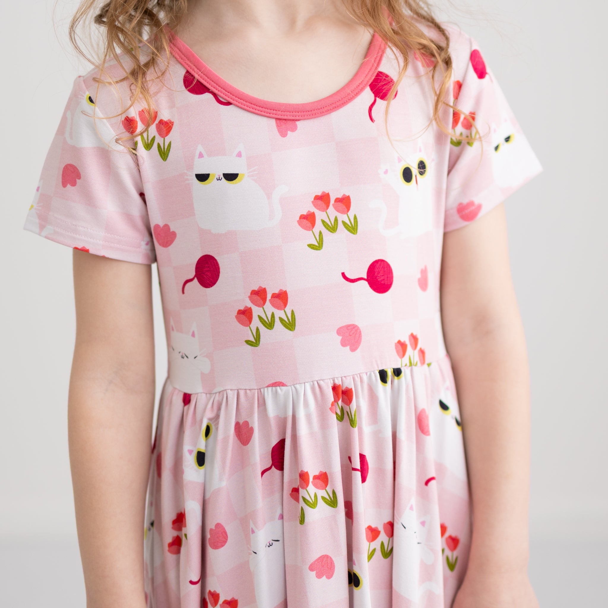A child wears a pink short-sleeved dress featuring a pattern of white cats, tulips, and yarn balls.
