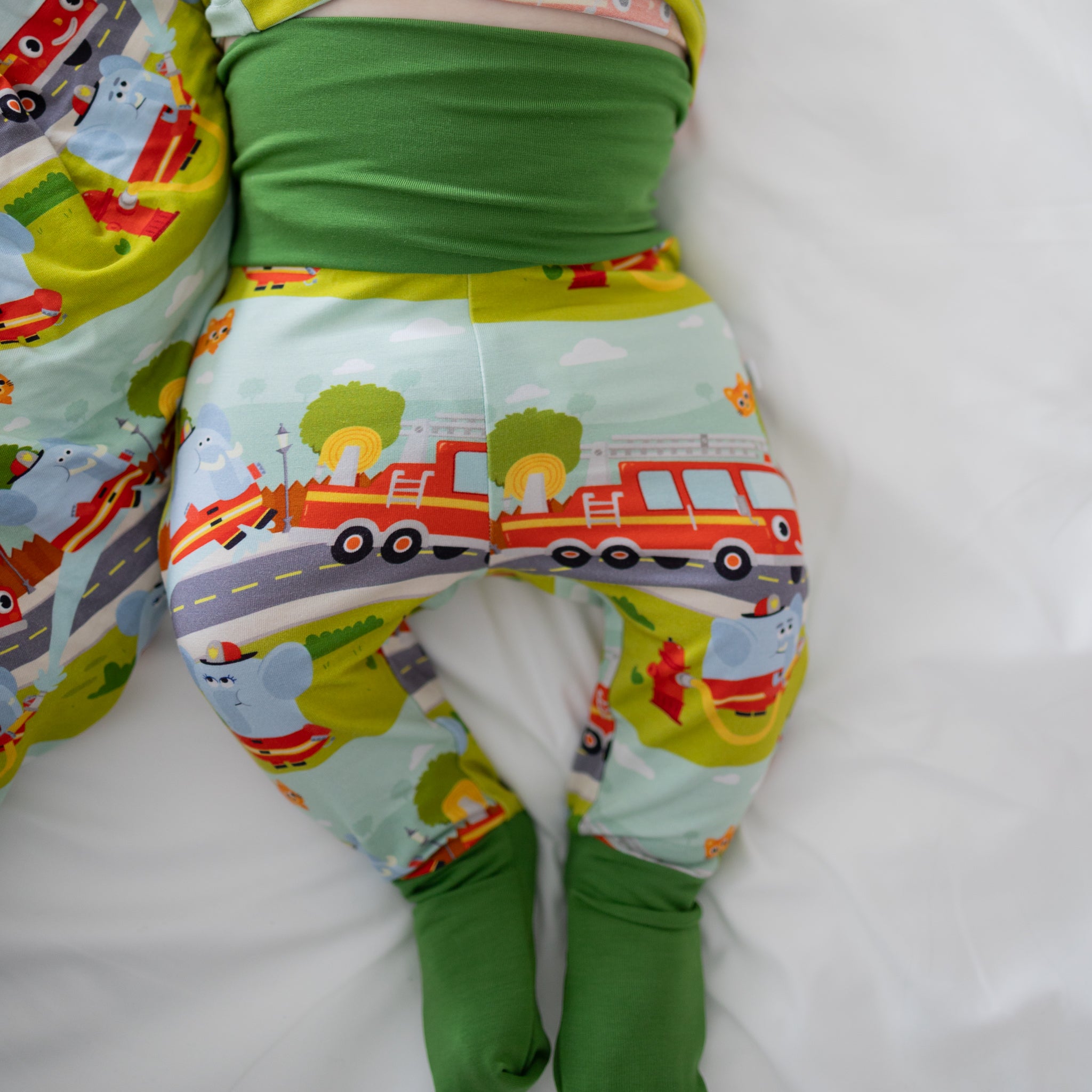 A baby wearing green-cuffed pants featuring a firetruck and elephant print on a white background.