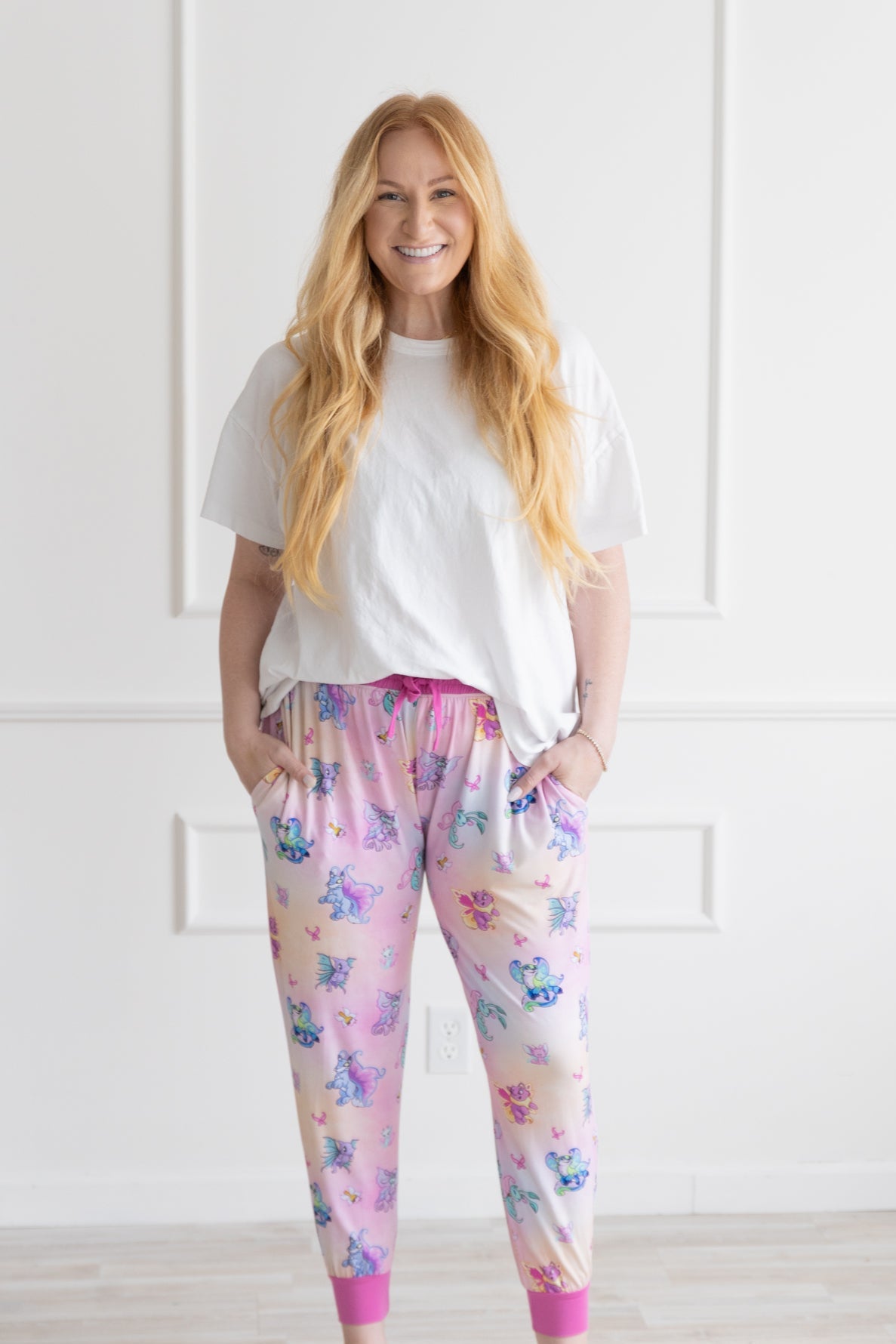 A woman wears pink and orange Neopets Faerie print jogger pants with a white t-shirt.