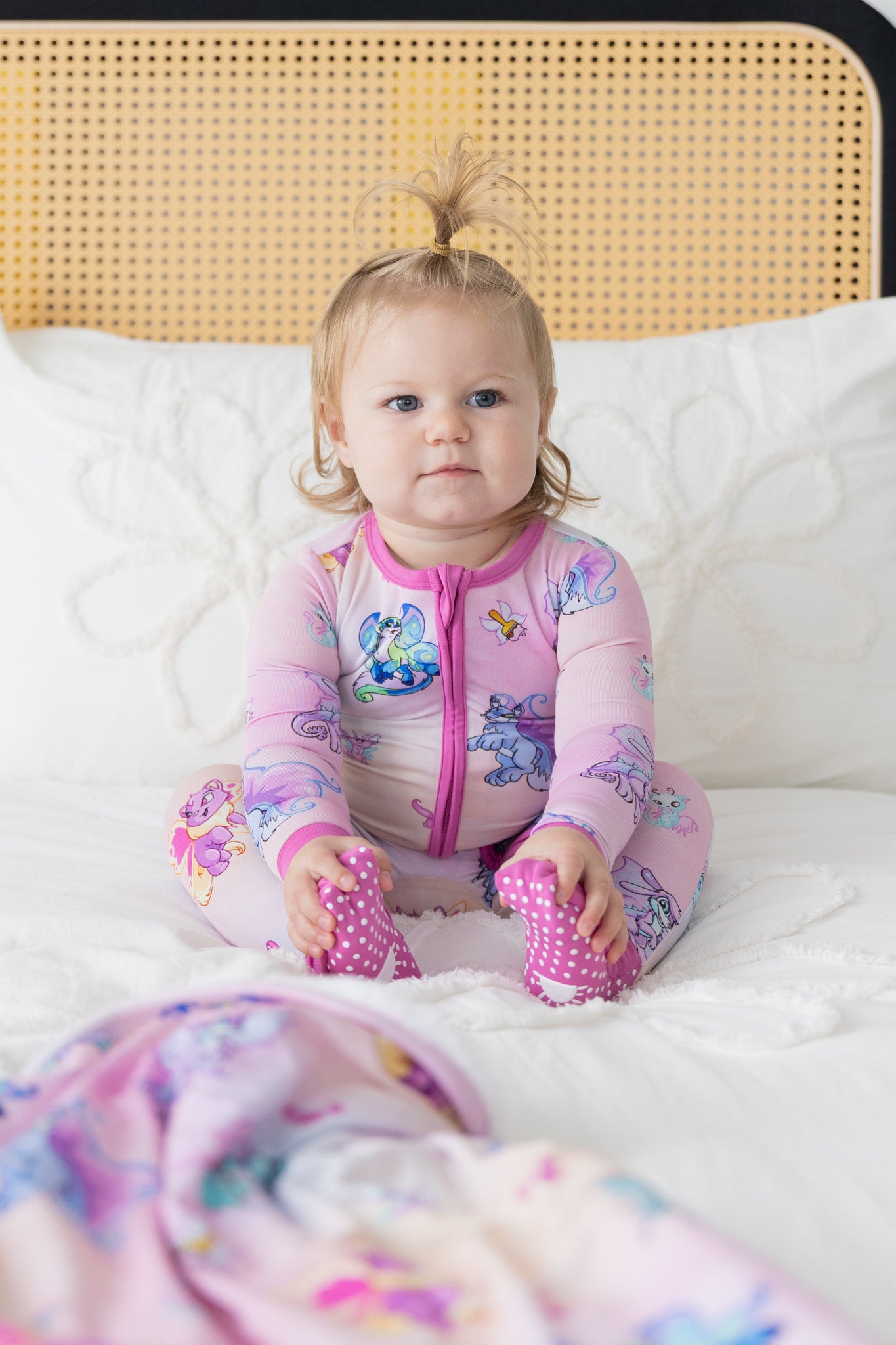 A baby sits on a bed wearing a pink Neopets Faerie print Tencel Modal zip-up pajama.