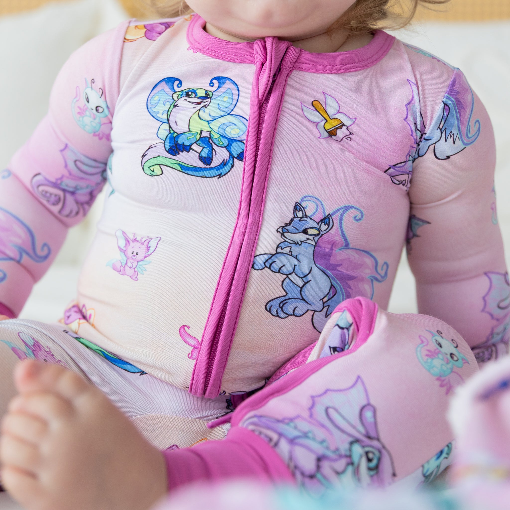 A baby wearing pink long-sleeve pajamas featuring a colorful Neopets Faerie print and a front zip.