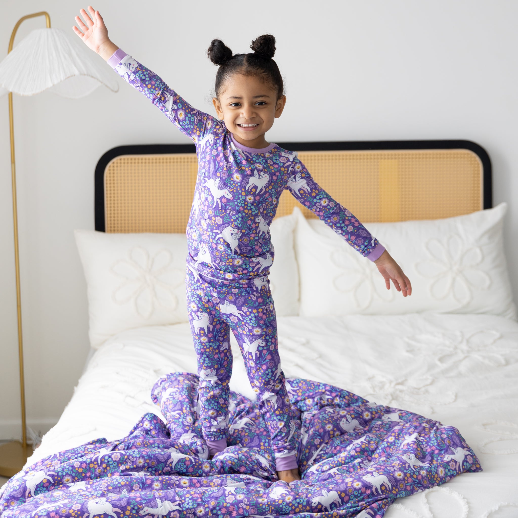 A young girl stands on a bed wearing purple unicorn-patterned pajamas made of Tencel Modal.