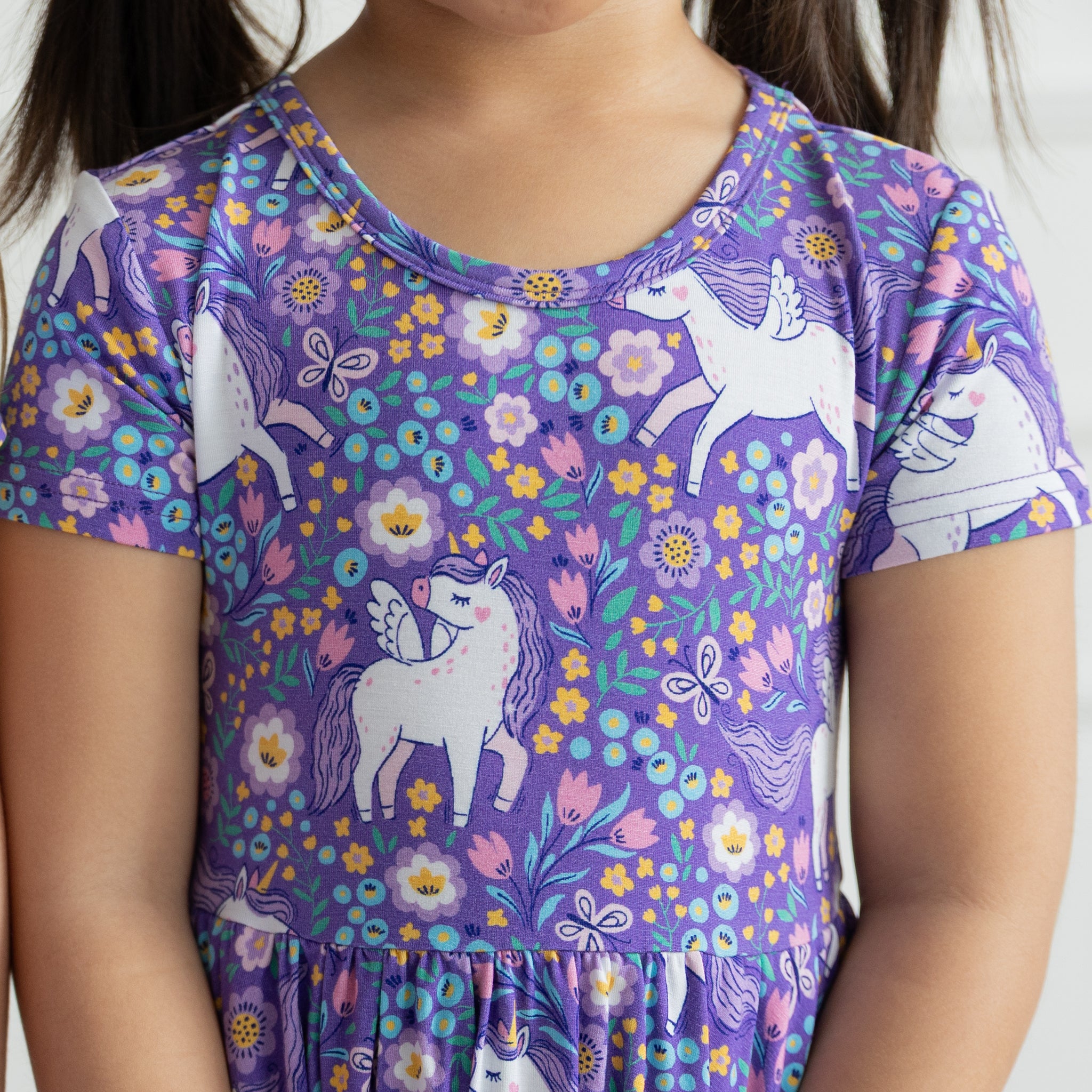 A child wears a purple short-sleeved dress featuring a pattern of white unicorns and flowers.