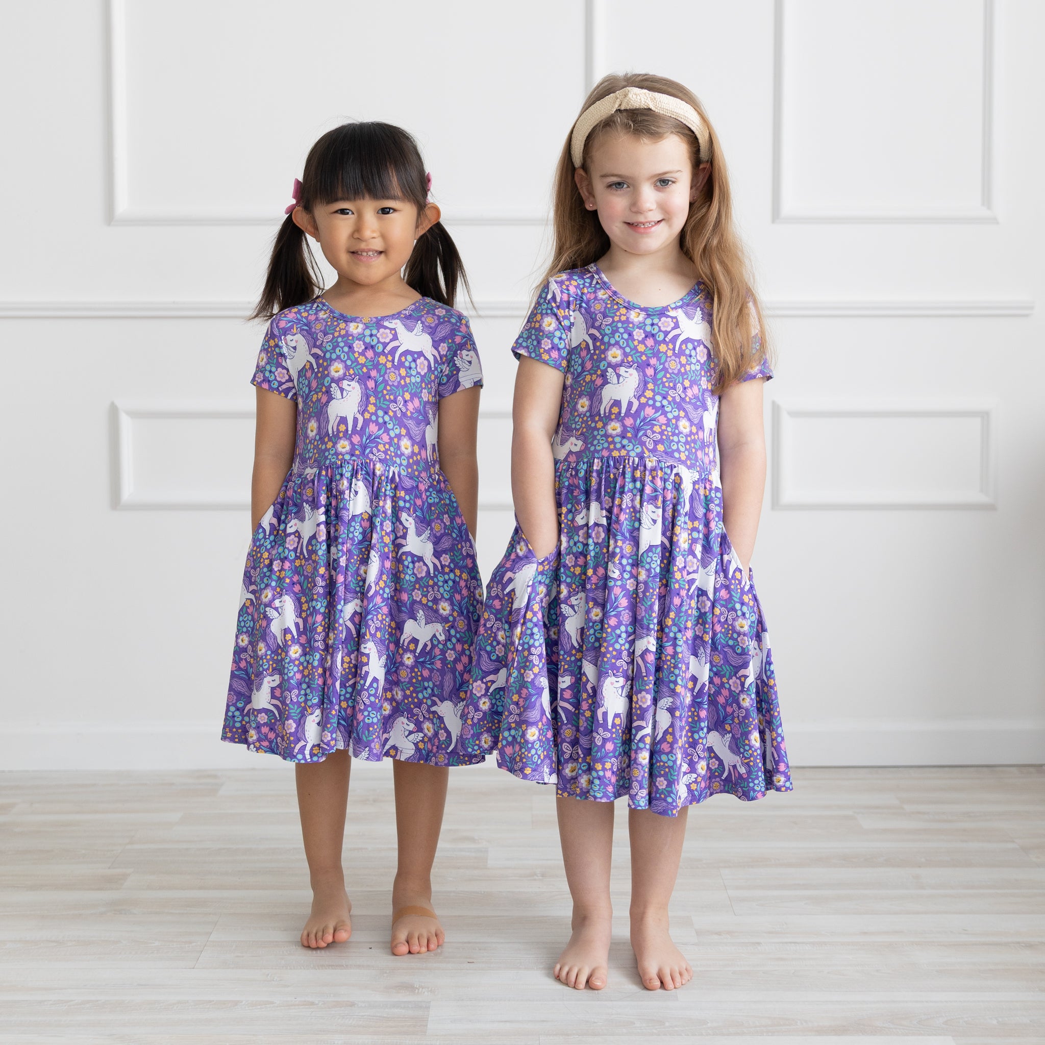 Two young girls stand side-by-side wearing matching purple short-sleeve dresses featuring a unicorn and floral print.