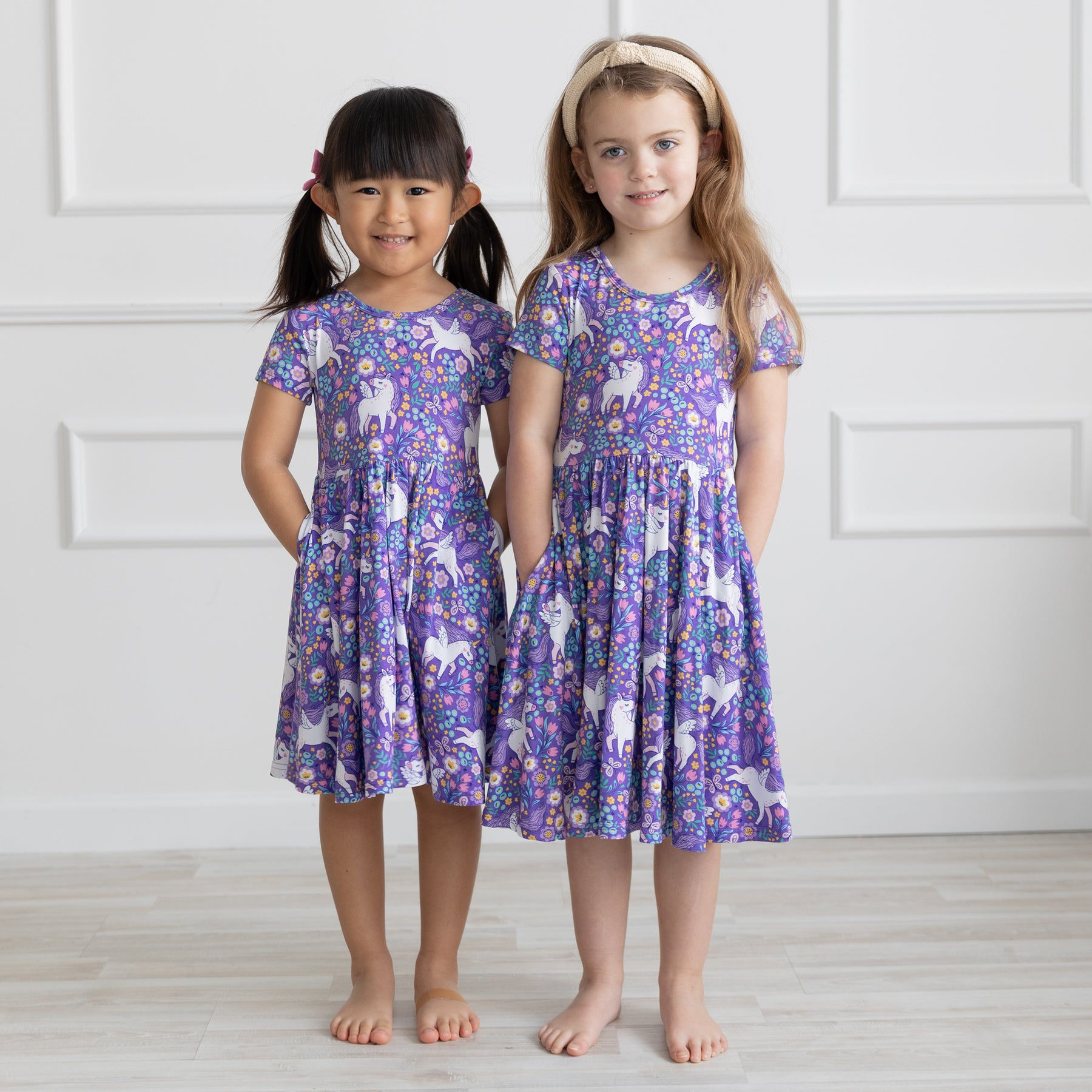 Two girls stand side-by-side wearing matching purple short-sleeve dresses with a unicorn print.