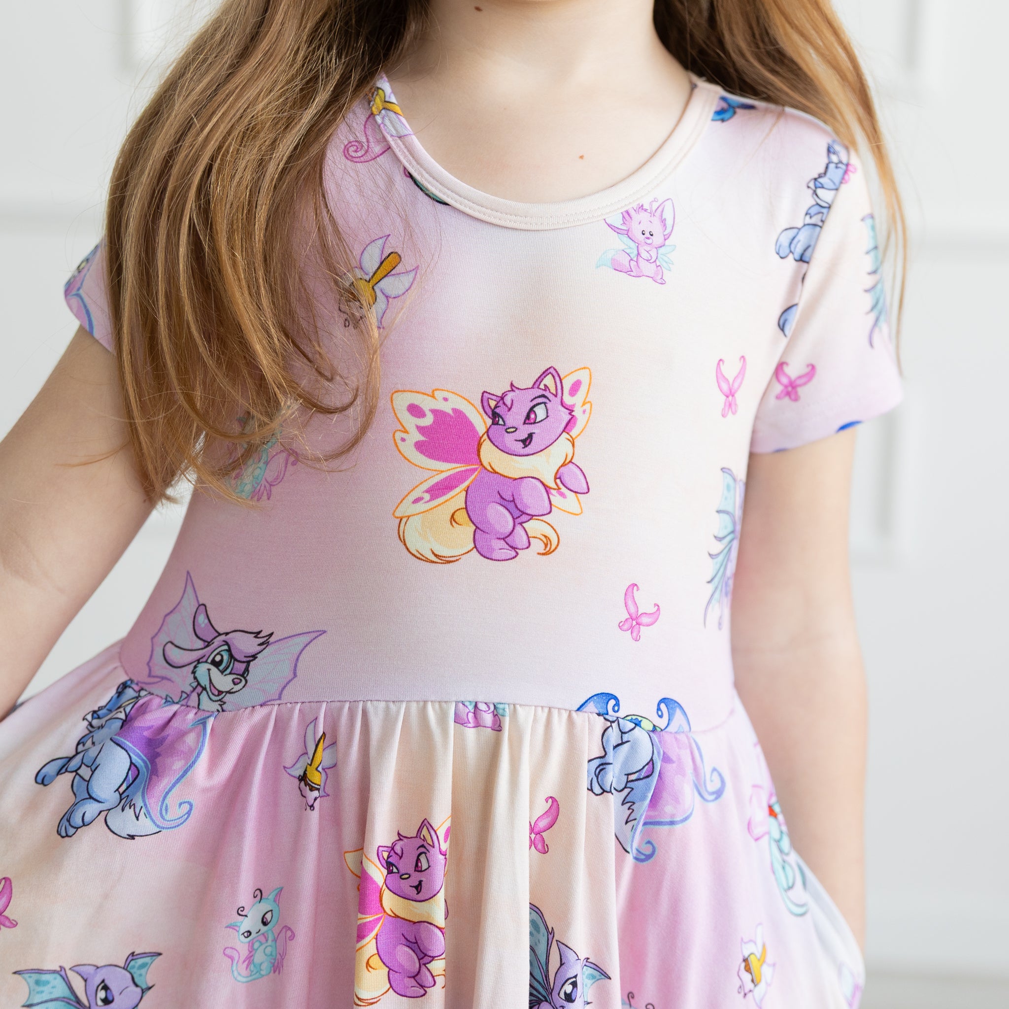 A child wears a light pink short-sleeved dress featuring a repeating pattern of Neopets faeries.