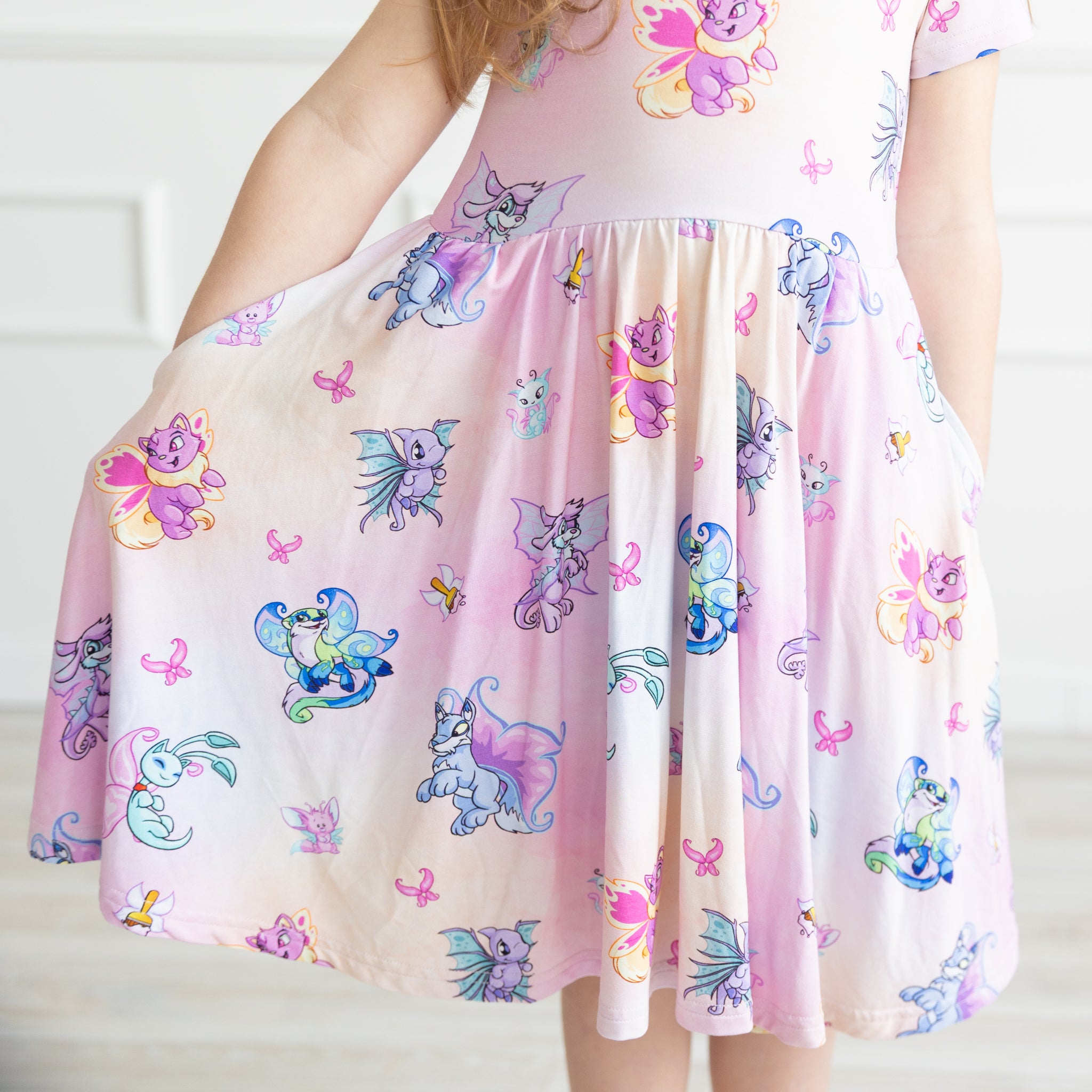 A child wears a pink short-sleeved dress featuring a colorful Neopets Faerie pattern and pockets.