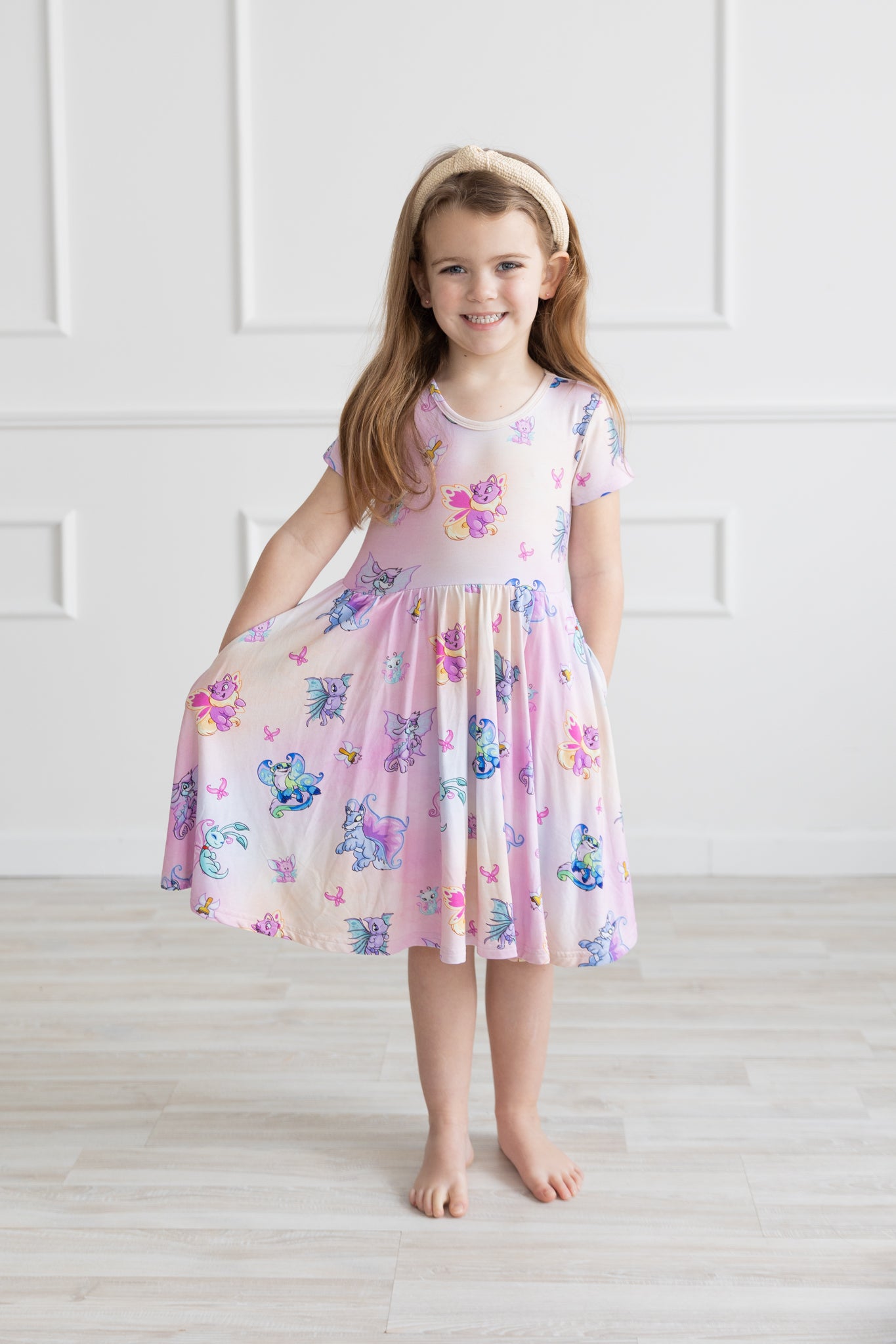 A young girl wears a pink and purple Neopets Faerie print dress with short sleeves and pockets.