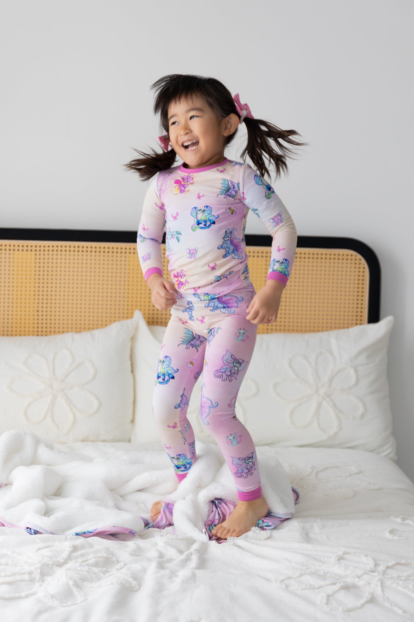 A child jumps on a bed wearing a pink long-sleeve pajama set featuring a Neopets Faerie print.
