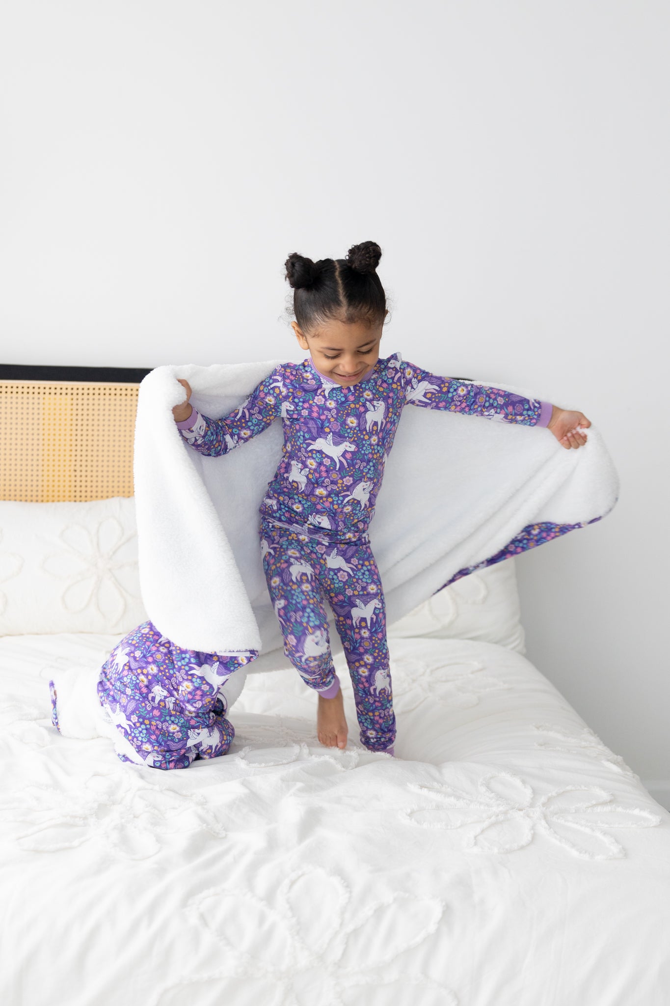A young girl wears a purple unicorn-patterned long-sleeve pajama set while holding a white blanket.