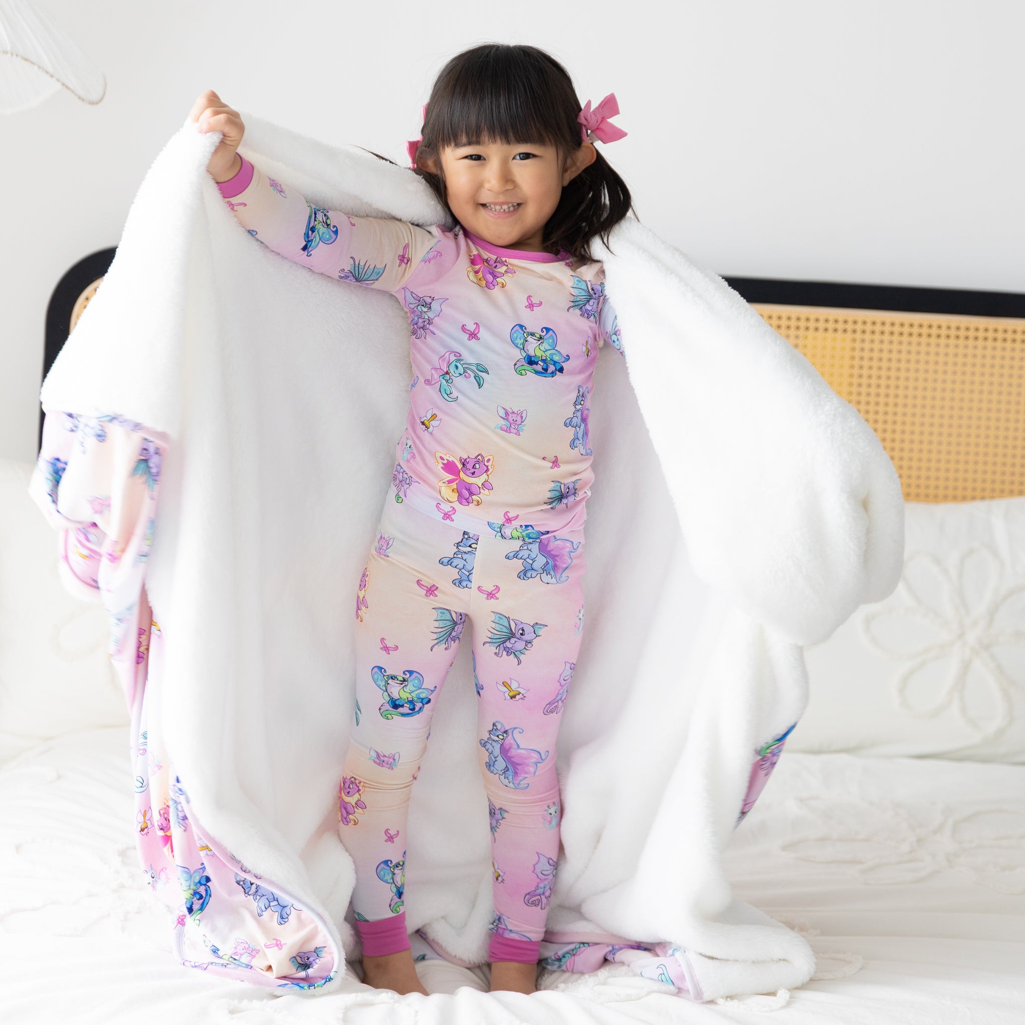 A young girl stands on a bed wearing a Neopets Faerie print pajama set and holding a matching blanket.