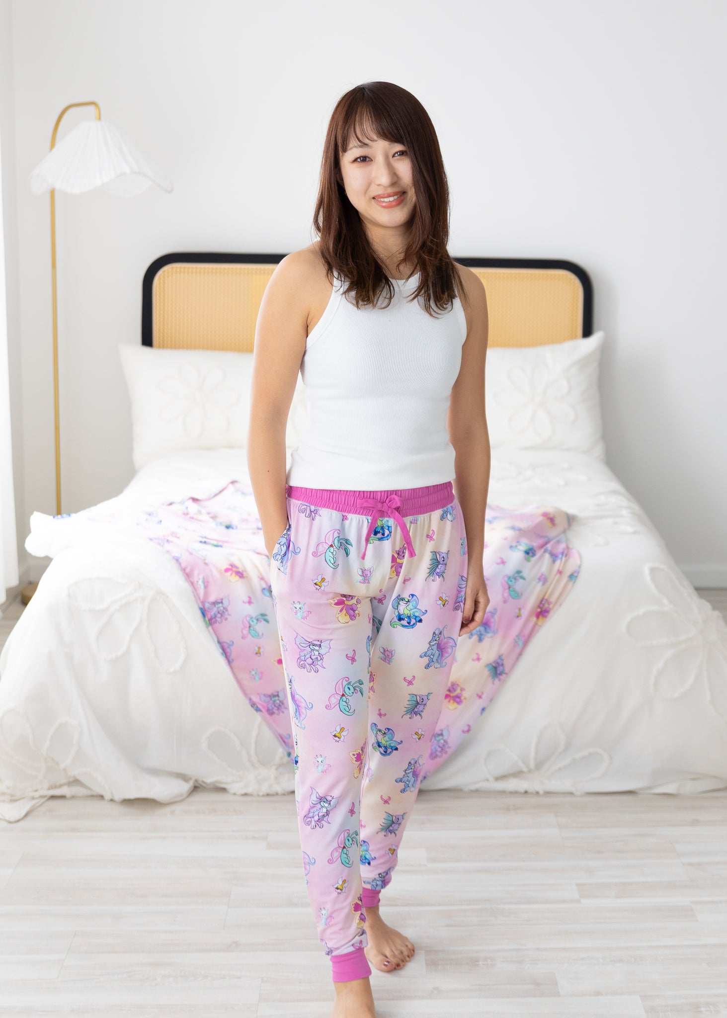 A woman wears pink Neopets Faerie print joggers with a white tank top in a bedroom.