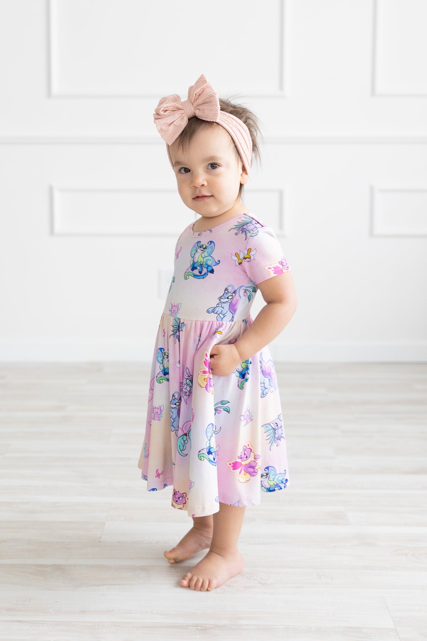 A toddler wears a short-sleeved pink and purple Neopets Faerie print dress with pockets.