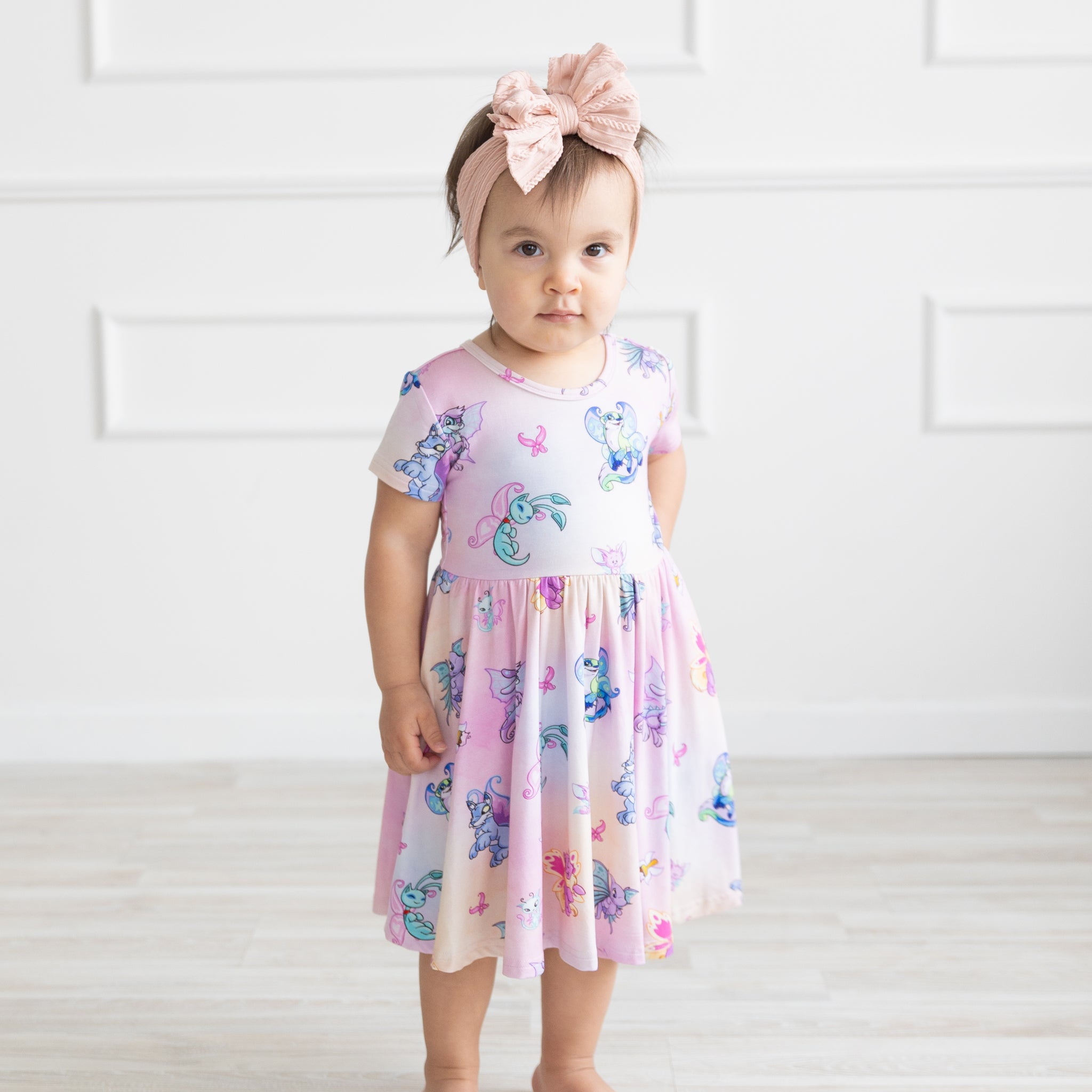 A toddler wears a short-sleeved pink dress featuring a colorful Neopets Faerie print.