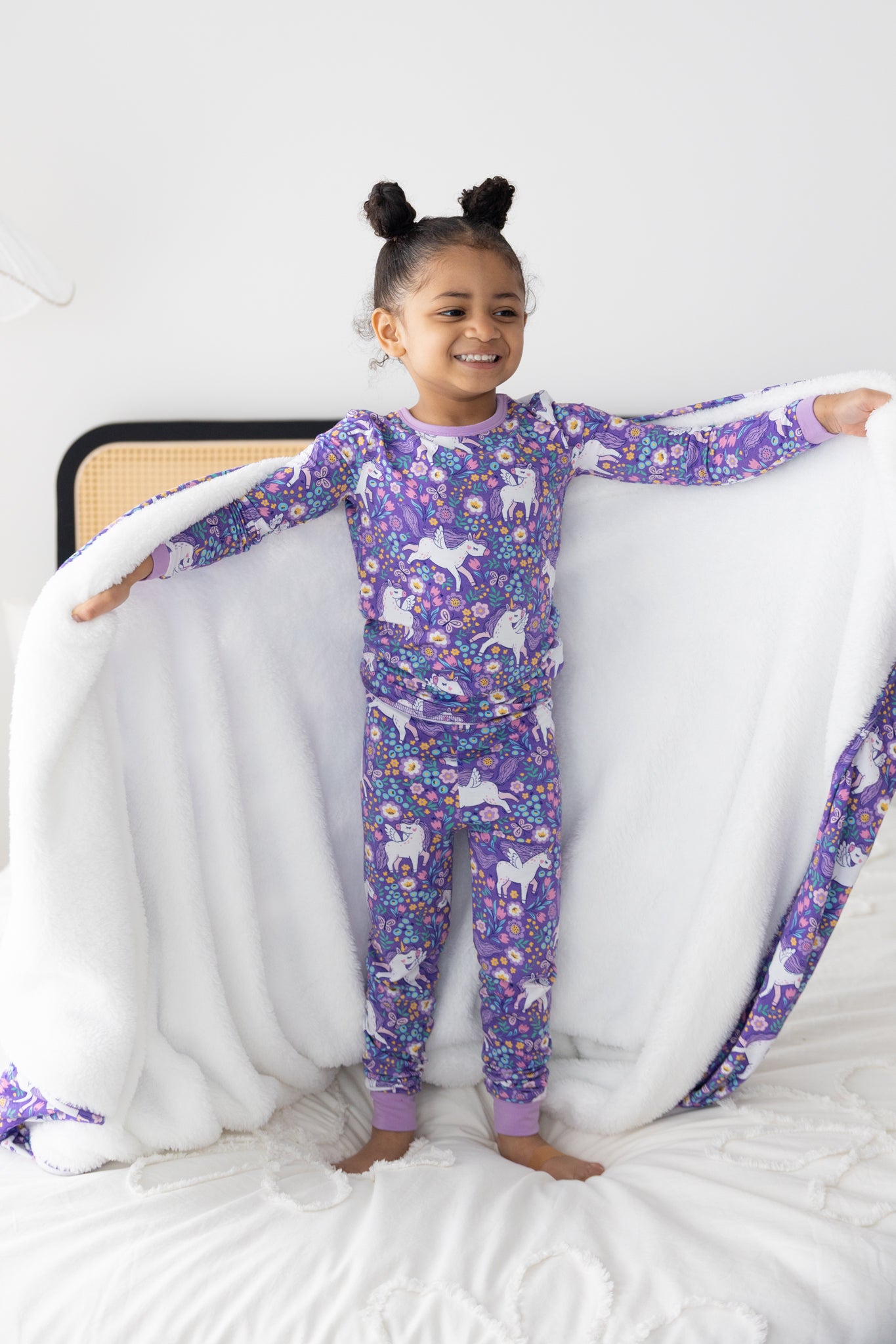A young girl stands on a bed wearing a purple unicorn-patterned long-sleeve pajama set.