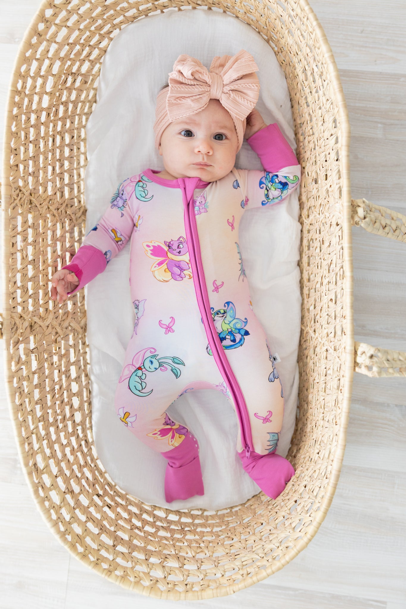 A baby wearing a pink Neopets Faerie print zip-up pajama with a matching bow headband.